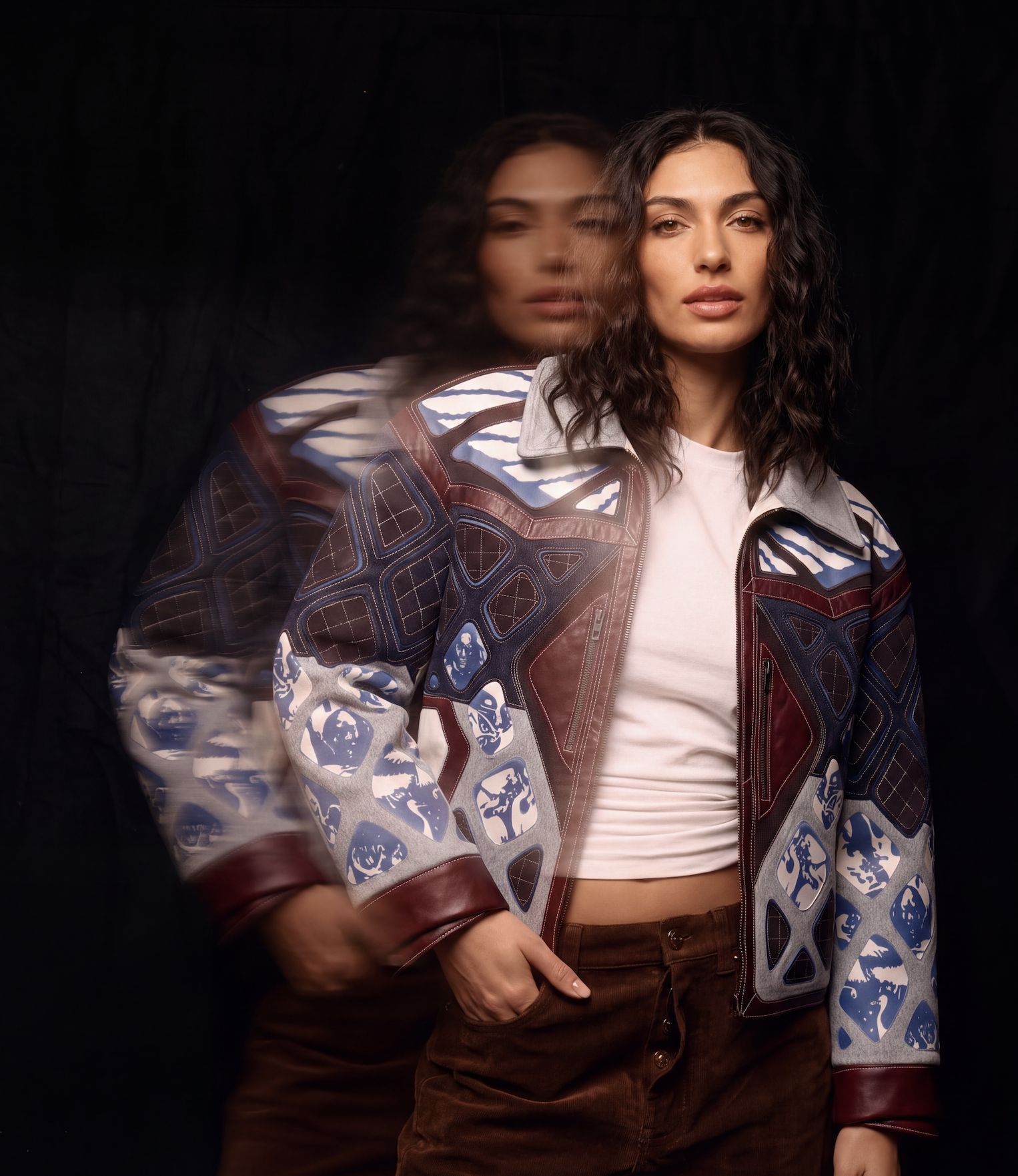 Long-exposure portrait of a model wearing The Egret Jacket, in burgundy leather and grey wool with blue printed panels, her movement trailing as a blur against a black backdrop
